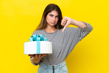 Young caucasian woman holding birthday cake isolated on yellow background showing thumb down with negative expression