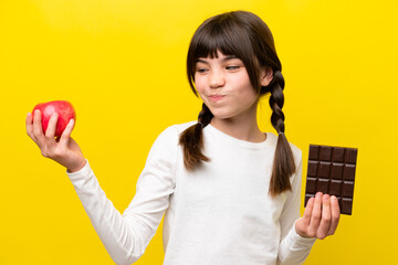 Little caucasian girl isolated on yellow background having doubts while taking a chocolate tablet...