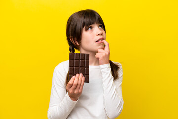 Little caucasian girl isolated on yellow background taking a chocolate tablet and having doubts