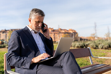 Concentrated senior businessman in formal wear sitting on bench and talking via smartphone while browsing laptop during remote work