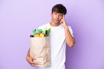 Young man holding a grocery shopping bag isolated on purple background thinking an idea