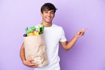 Young man holding a grocery shopping bag isolated on purple background pointing finger to the side