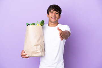 Young man holding a grocery shopping bag isolated on purple background shaking hands for closing a good deal