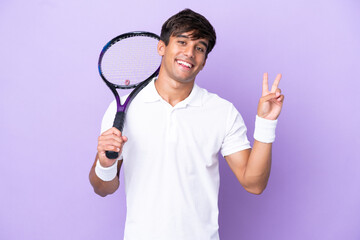 Handsome young tennis player man isolated on ocher background smiling and showing victory sign