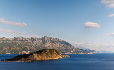Island of St. Nicholas with vegetation on shores in the Adriatic Sea against the backdrop of coastal towns, the Montenegrin mountains and clear sky
