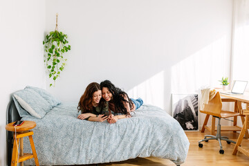 Young women friends having fun using mobile phone while relaxing together on bedroom