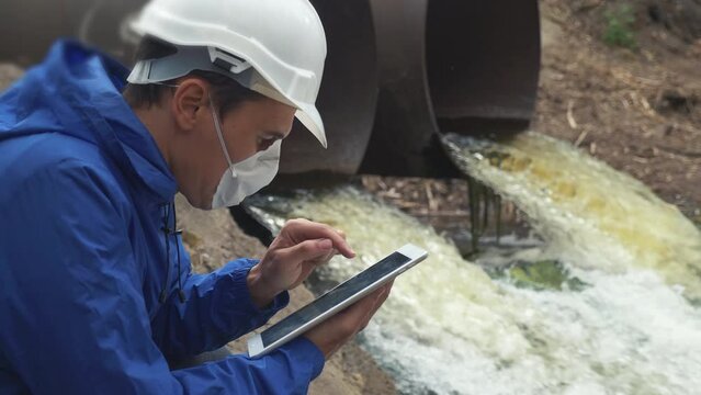 environmental pollution lifestyle radiation waste concept. man chemist scientist in a respirator mask studies water pollution digital tablet and an white helmet stands next to the waste pipes
