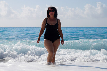 a curvaceous woman in a black bathing suit and in sunglasses stands in the sea, raising her hands