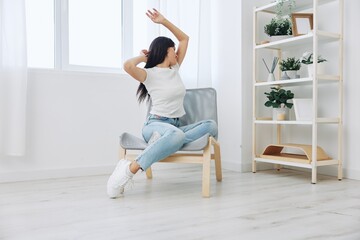 Woman sitting in a chair listening to music and dancing with wireless headphones at home in jeans and a white T-shirt, fall lifestyle comfort