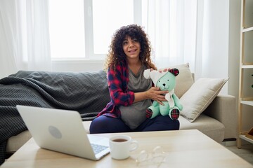 A pregnant woman sits on a couch with a rabbit toy and looks into a laptop. Lifestyle of a pregnant woman, preparation for childbirth, last month of pregnancy