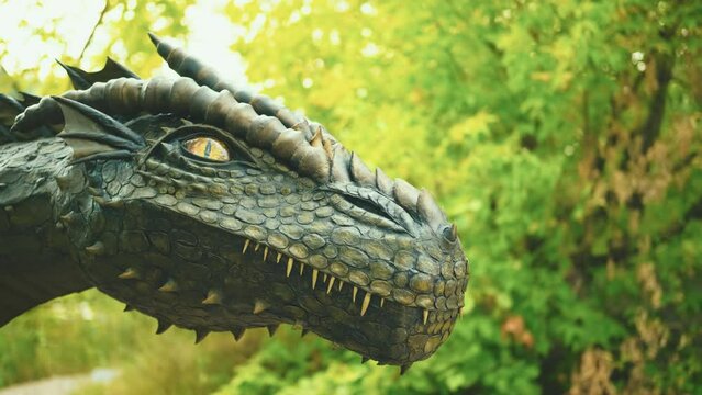 Fantasy portrait real dragon head close-up sharp teeth beautiful eyes muzzle in dark gray-green scales, spikes. Creative big dinosaur toy of Jurassic period. Background summer nature trees foliage. 4k