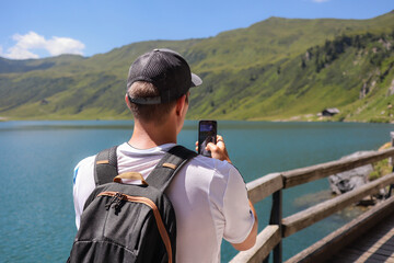 Obraz premium Caucasian Man from Behind with Smartphone in European Nature. Young Adult with Backpack and Cap Takes Picture with Phone of Scenic Outdoor Scene of Alpine Lake during Summer Sunny Day in Austria.