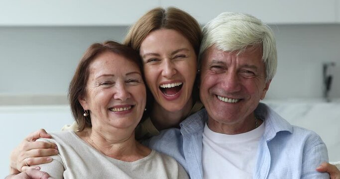 Cheerful excited loving adult daughter hugging couple of senior parents from behind, laughing, looking at camera with toothy smile. Older mom and dad and grownup kid head shot portrait