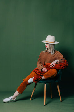 Stylish blond hair woman in hat nad orange pants sits in chair on green backgorund in studio