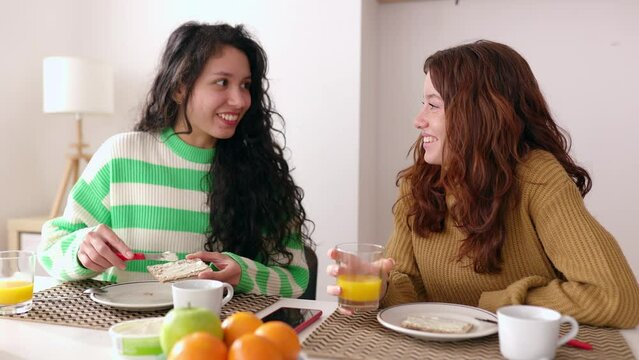 Young diverse female friends talking while having breakfast in the living room at home. Domestic lifestyle concept