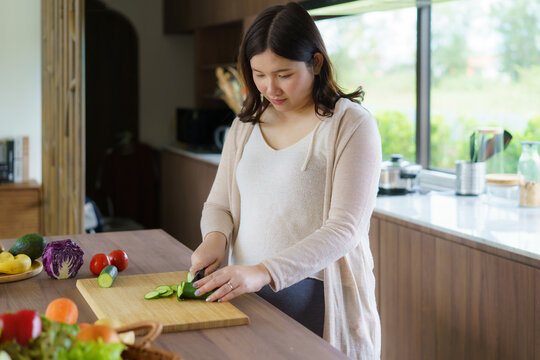Pregnant Asian woman cutting cucumber for fresh green salad, female prepares tasty organic dinner at home, healthy nutrition for future mother