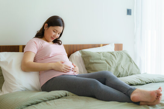 Pregnancy, people rest and expectation concept - Asian smiling happy pregnant woman sitting in bed and touching her belly at home.
