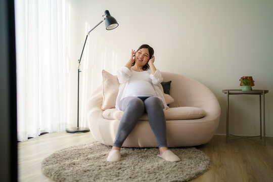 Portrait of pregnant Happy Asian woman sitting on sofa at home and using smart phone for listening music in headphones.