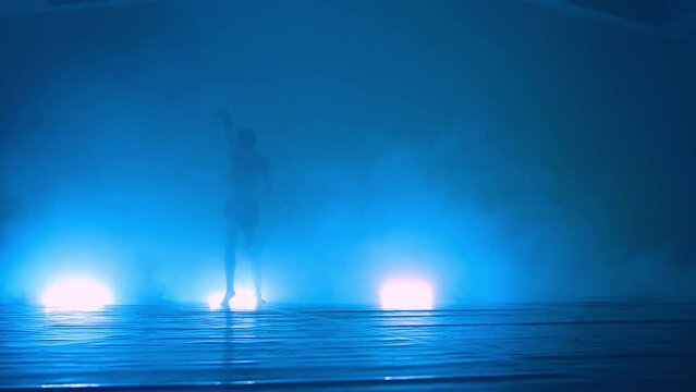 A young male ballet dancer with naked torso performs dance moves against a blue background, with a light of lights smoke