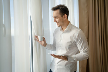 Naklejka na ściany i meble man wear white shirt hold cup of coffee and digital tablet at window