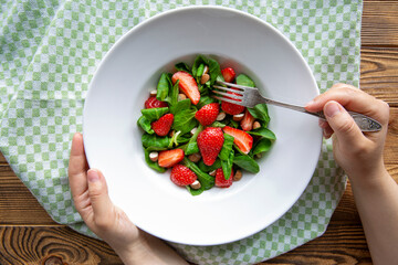 Delicious fruit summer salad, female hands hold plate and fork, rustic wooden background, top view. 