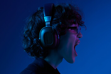 Teenage man wearing headphones listening to music and dancing and singing open mouth with glasses, hipster lifestyle, portrait blue background, neon light, style and trends, mixed light, copy space