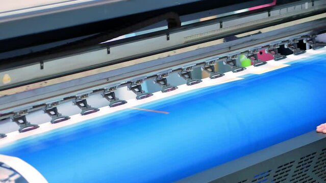 The printhead of a large-format inkjet printer moves along the banner material, the blue background of the chrome key is applied with ink. Production of advertising products. Close-up.