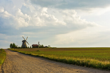 Fototapeta premium Dirt road to the lonely windmill and farmhouse located among the field during sunset