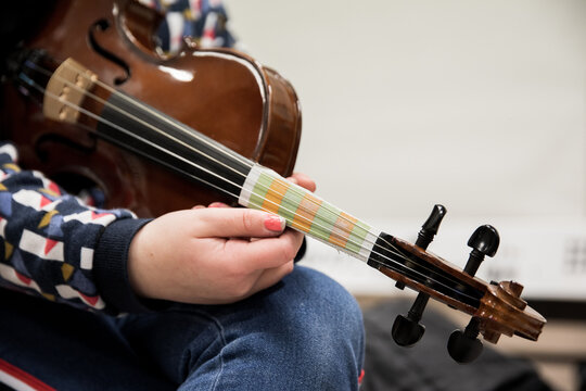 Photographie des mains d'une enfant aux ongles vernis tenant son violon d'apprentissage, l'emplacement des notes de musique sont color&eacute;es sur un papier positionn&eacute; sur le manche du violon.