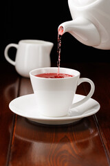 Red tea is poured from the teapot into a cup. In the background is a milk jug. Side view. Selective focus.