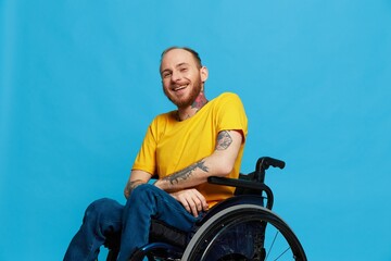 A man in a wheelchair smile looks at the camera in a t-shirt with tattoos on his arms sits on a blue studio background, a full life, a real person