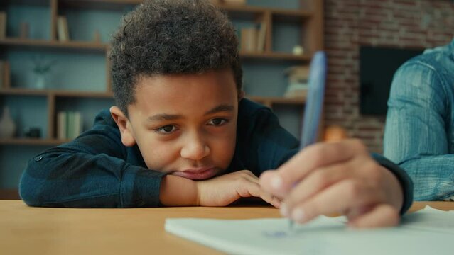 Tired exhausted sad upset fatigued little African American ethnic child boy kid son pupil schoolboy schoolchild lying on table desk writing homework boring class lesson drawing in notebook near father