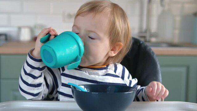 One year old hungry baby girl in striped casual clothes sits at white table in gray highchair and drinks herself from bottle. Blurred dining room background. Healthy eating for kids. Child's nutrition
