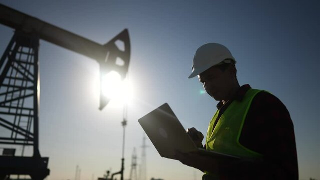 oil business. a worker works next to an oil pump lifestyle holding a laptop. industry business oil and gas concept. engineer studying the level of oil production on a laptop silhouette at sunset