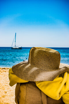 Un chapeau en paille est pos&eacute; sur une chaise sur une plage  en &eacute;t&eacute; devant une mer bleu turquoise , on devine un voilier au loin