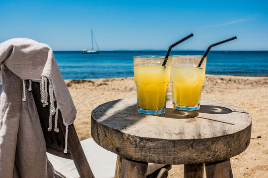 Des cocktails de fruits sont pos&eacute;s sur une jolie table en bois sur une plage magnifique en &eacute;t&eacute;, on devine un voilier mouill&eacute; au large .
