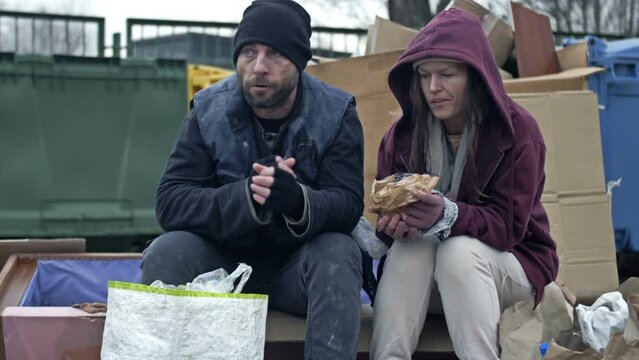 Two dirty and poorly dressed homeless people, a man and a woman, sit by a pile of rubbish and eat something. Young woman is sick and coughing.