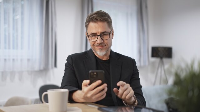 Businessman at home, using phone, checking newsfeed. Home office, browsing internet. Portrait of happy, mature age, middle age, mid adult man in 50s, smiling.