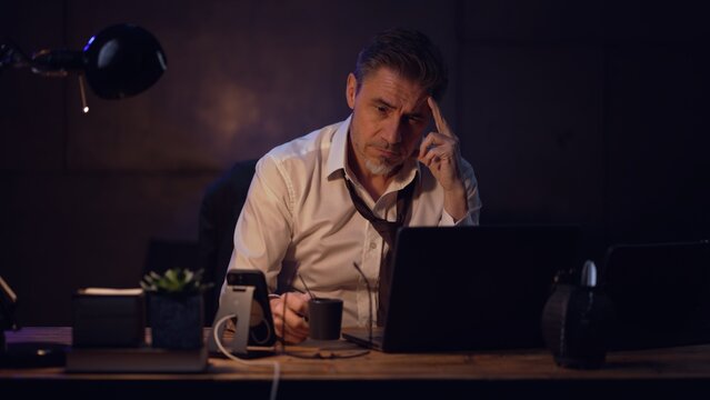 Businessman sitting at desk with laptop computer, working late in office, tired. Entrepreneur thinking, solving problem. Older, middle aged, mid adult, man in his 40s or 50s in shirt and tie.