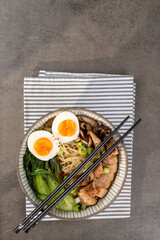 Asian noodle soup, ramen with chicken breast, vegetables, mushrooms and egg in the bowl on a striped napkin and gray concrete background. Top view, healthy eating concept.