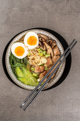 Asian noodle soup, ramen with chicken breast, vegetables, mushrooms and egg in bowl on a black plate and gray concrete background. Top view, healthy eating concept