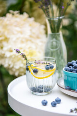 A glass with delicious homemade lemonade made of fresh blueberries, lemon and lavender on white round table on backyard in summer day and white blooming hydrangea flowers on background.