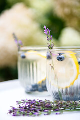 Two sweaty glasses with cold delicious homemade lemonade made of fresh blueberries, lemon and lavender on white table on backyard in summer day and blooming hydrangea flowers on background.