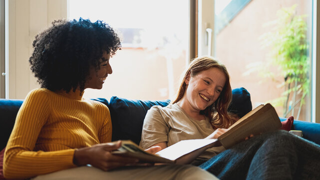 two young women at home, a couple of lesbian sitting in sofà in living room, reading books and having moments of relax, smiling african woman looking her girlfriend reading