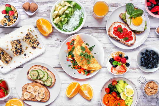 Healthy breakfast food table scene. Overhead view on a white wood background. Omelette, nutritious bowl, toasts, granola bars, smoothie bowl, yogurts and fruits.