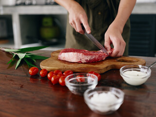 Woman with knife in hand cutting fresh steak meat for roasting in kitchen with salt pepper and other spices on table, red cherry tomatoes and herbs, preparing dinner.