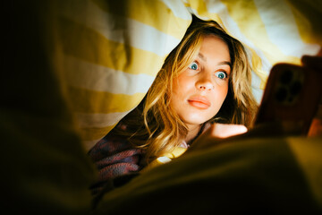 Young amazed caucasian woman using smartphone, lies under a blanket on a bed at home in the evening.