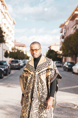 A dark-skinned man posing in the middle of the street dressed in a poncho with Aztec drawings and sunglasses, the black male is facing the camera.