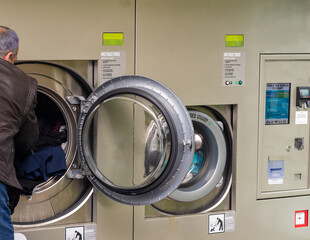 senior male, wearing jeans and brown jacket, using self-service laundry, inserting dirty clothes...
