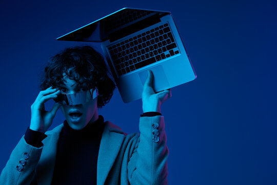 Portrait of a stylish male hacker with a laptop and futuristic glasses in blue light, cyber security, technology and education, neon color, freelancer online work